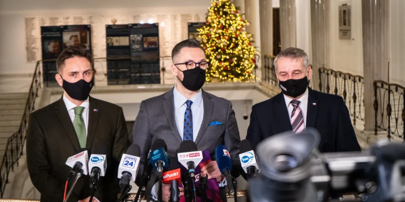 Trzej mężczyźni w eleganckich strojach, noszący maseczki, stoją przed mikrofonami w dekorowanej bożonarodzeniowo przestrzeni.