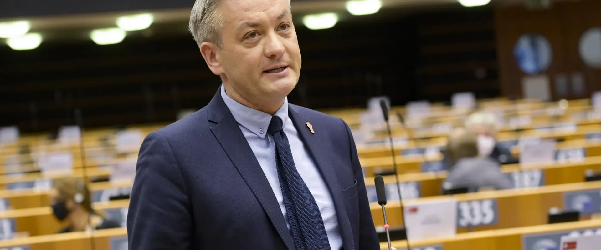 Mężczyzna w garniturze przemawia w sali obrad, otoczony przez inne osoby siedzące w biurkach.