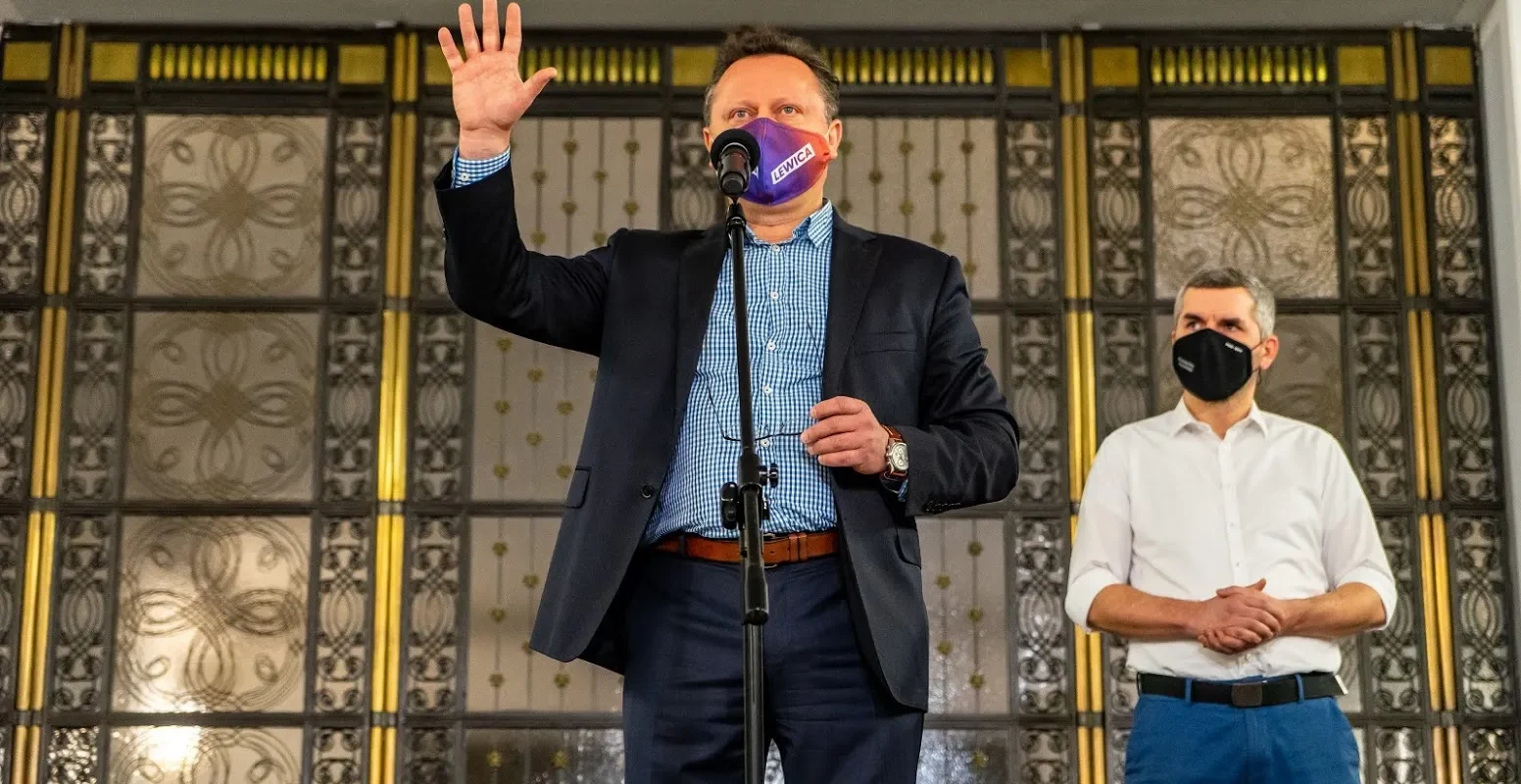 Trzech mężczyzn w maseczkach na scenie przed dekoracyjnymi drzwiami; centralny mężczyzna w garniturze z fioletową maską z napisem LEWICA unosi dłoń przy mikrofonie, a drugi stoi obok z założonymi rękami.