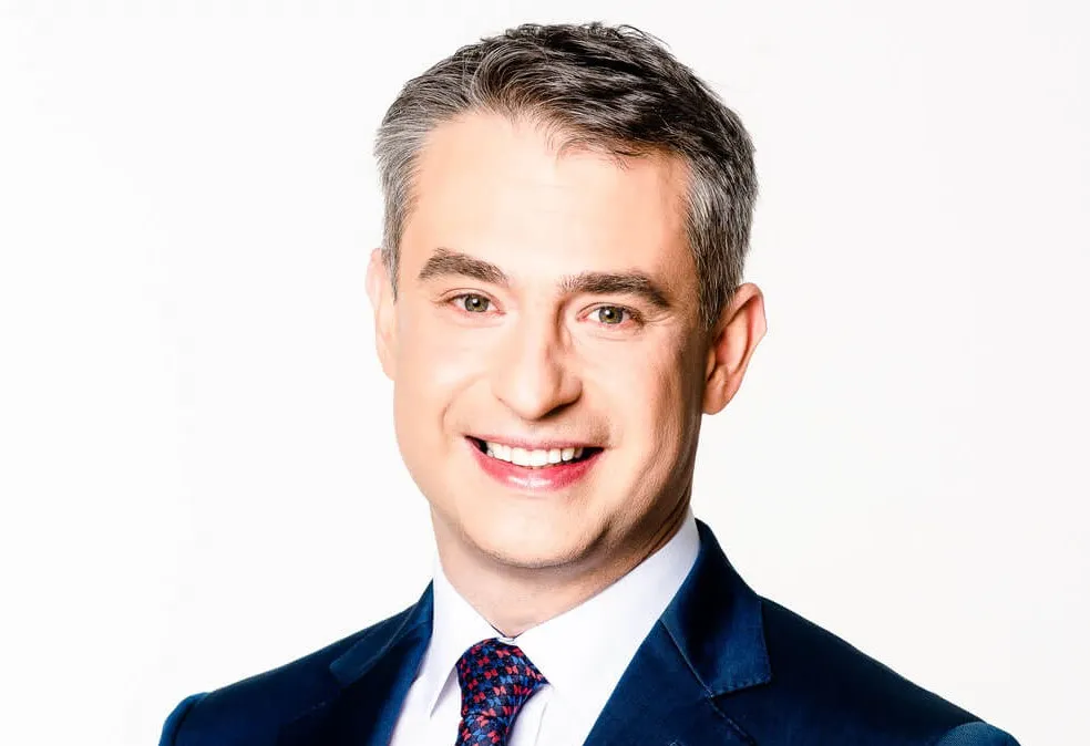 Portret uśmiechniętego mężczyzny w granatowym garniturze na białym tle.