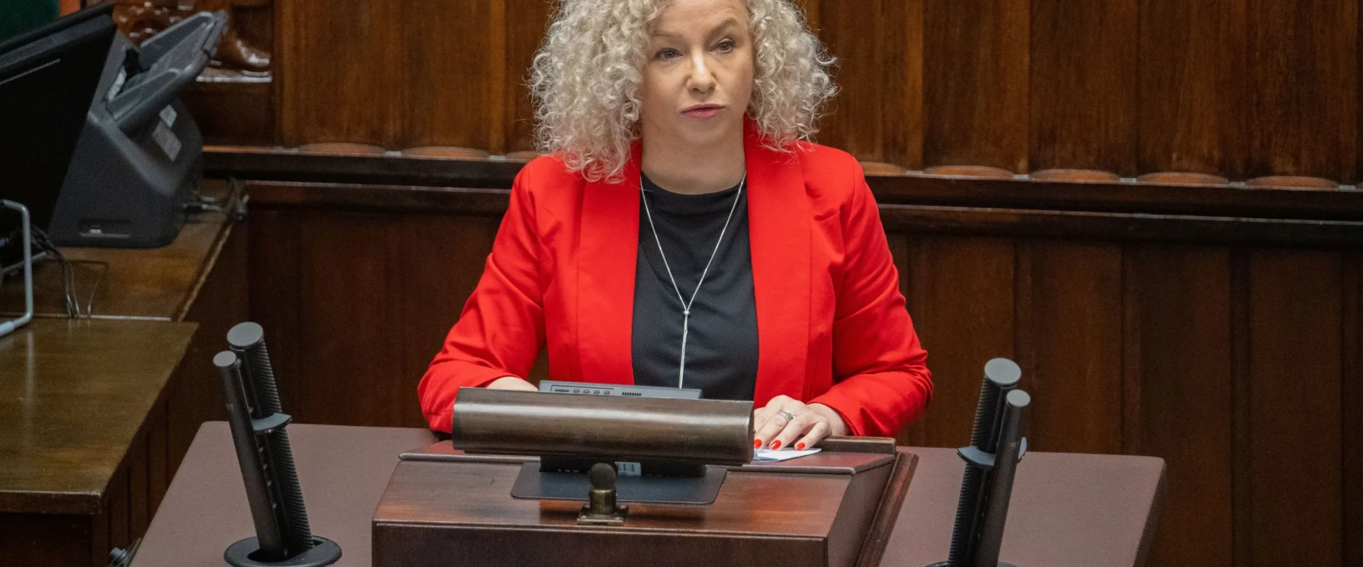 Kobieta o kręconych siwych włosach, ubrana w czerwoną marynarkę, siedzi za mównicą w sali obrad podczas debaty o aborcji.