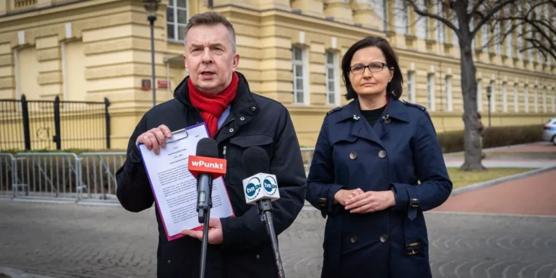 Mężczyzna przemawia do mediów, trzyma dokumenty i mikrofon, stojąc obok kobiety podczas konferencji prasowej na tle żółtego budynku; temat dotyczy inwestycji w odnawialne źródła energii zamiast rosyjskich surowców.