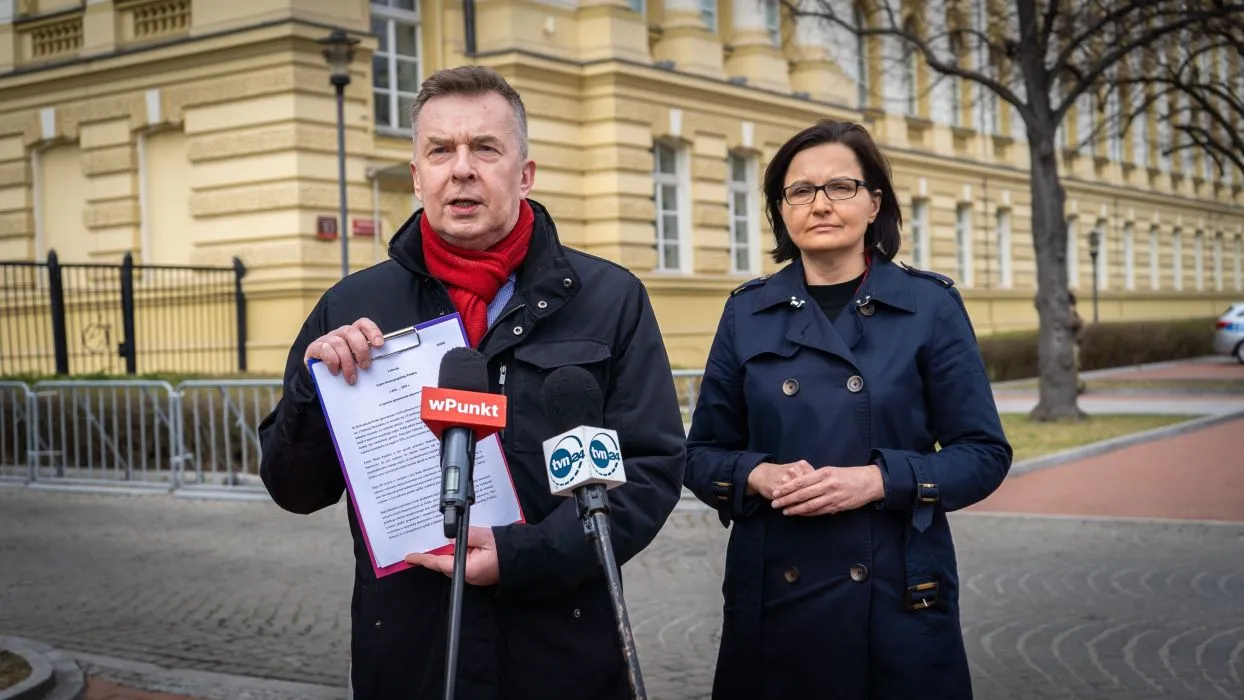 Mężczyzna przemawia do mediów, trzyma dokumenty i mikrofon, stojąc obok kobiety podczas konferencji prasowej na tle żółtego budynku; temat dotyczy inwestycji w odnawialne źródła energii zamiast rosyjskich surowców.