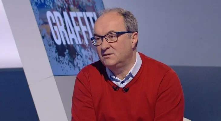 Osoba w czerwonym swetrze i okularach siedzi w studiu telewizyjnym z mikrofonami, przed dużym ekranem z napisem "GRAFFITI".