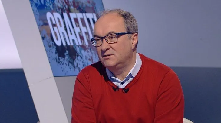 Osoba w czerwonym swetrze i okularach siedzi w studiu telewizyjnym z mikrofonami, przed dużym ekranem z napisem "GRAFFITI".