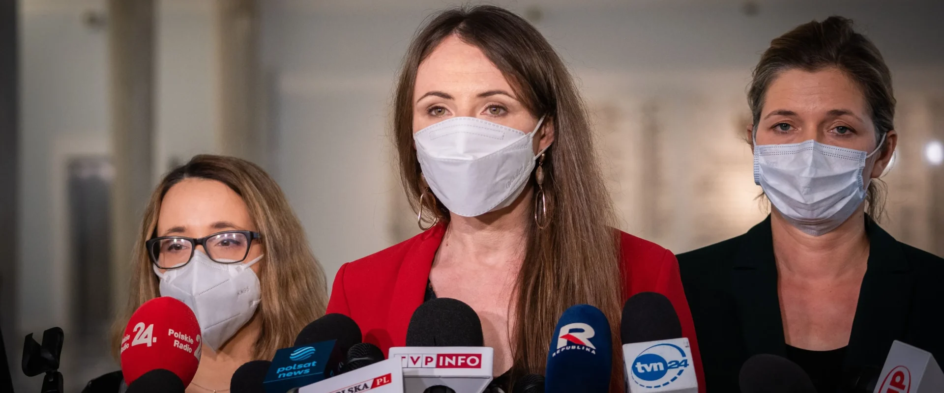 Na zdjęciu widać trzy kobiety stojące przed mikrofonami, każda w maseczce ochronnej, podczas konferencji prasowej.