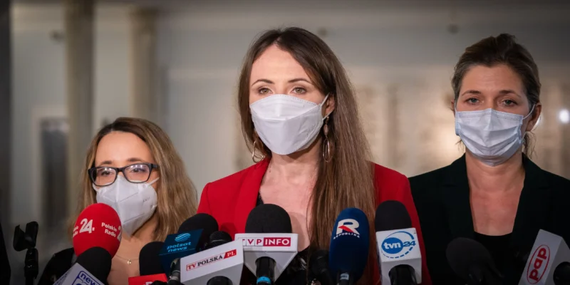 Na zdjęciu widać trzy kobiety stojące przed mikrofonami, każda w maseczce ochronnej, podczas konferencji prasowej.