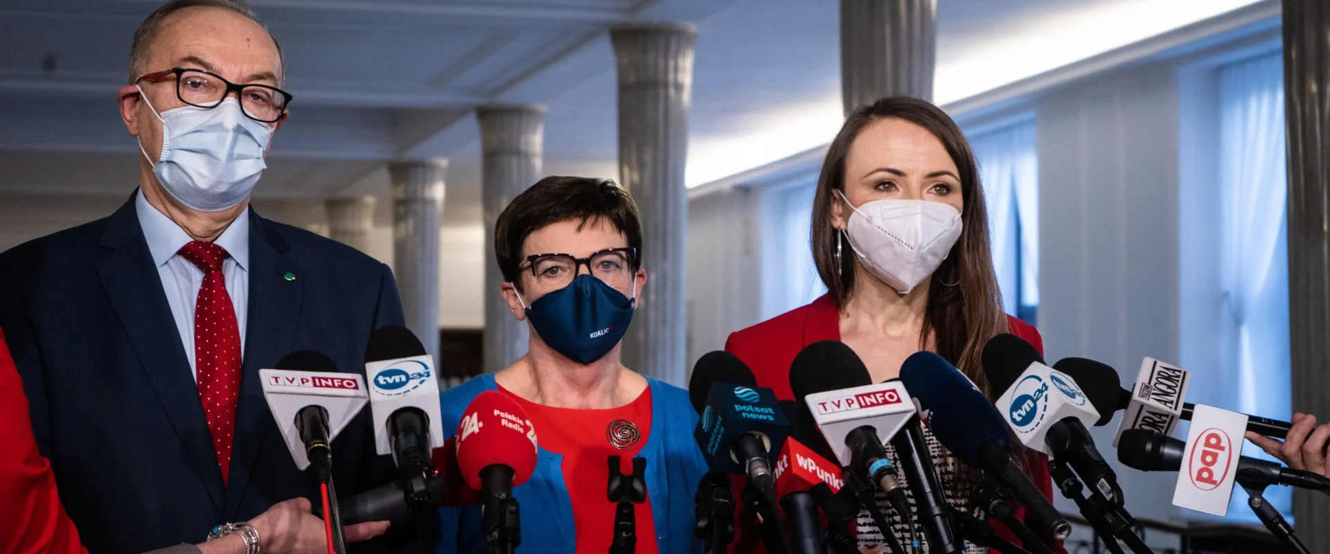 Trzy osoby w maseczkach i garniturach stoją przed licznymi mikrofonami podczas konferencji prasowej w korytarzu z kolumnami.