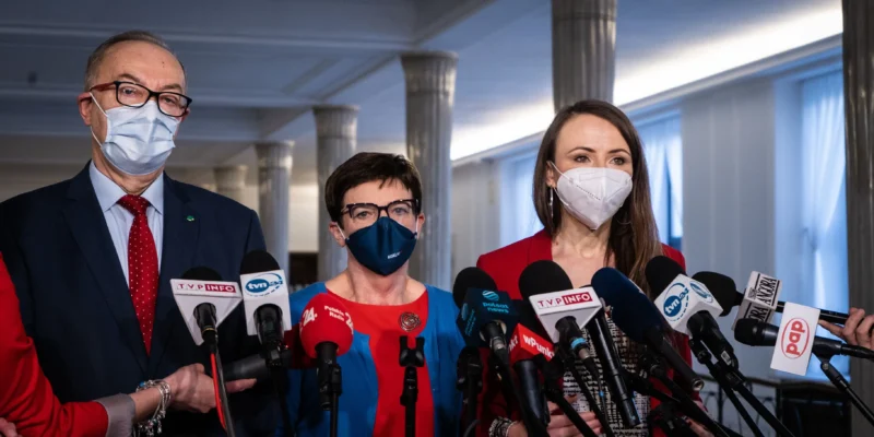 Trzy osoby w maseczkach i garniturach stoją przed licznymi mikrofonami podczas konferencji prasowej w korytarzu z kolumnami.