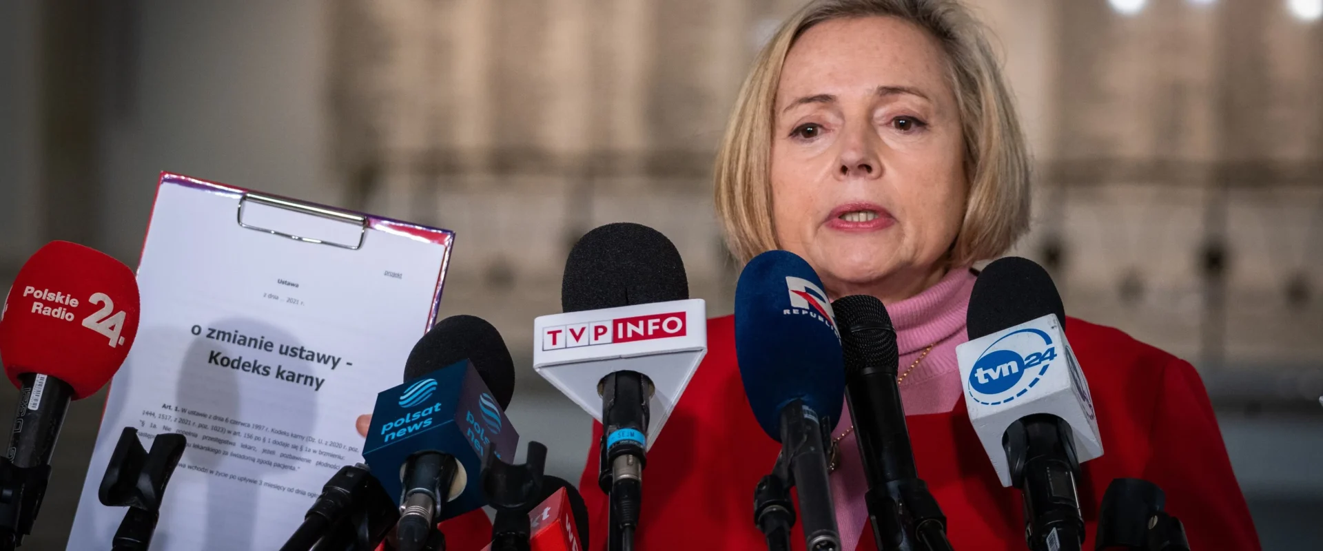 Na zdjęciu widoczna jest kobieta w czerwonym ubraniu, stojąca przed mikrofonami, podczas konferencji prasowej na temat zmiany ustawy dotyczącej kodeksu karnego.