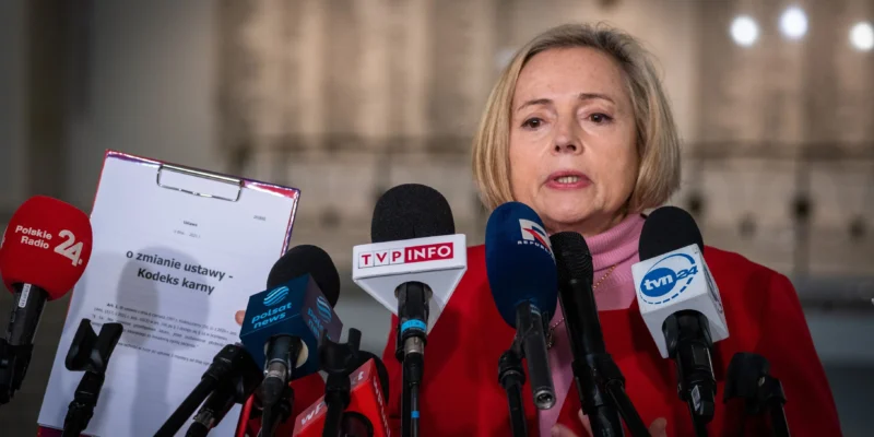Na zdjęciu widoczna jest kobieta w czerwonym ubraniu, stojąca przed mikrofonami, podczas konferencji prasowej na temat zmiany ustawy dotyczącej kodeksu karnego.