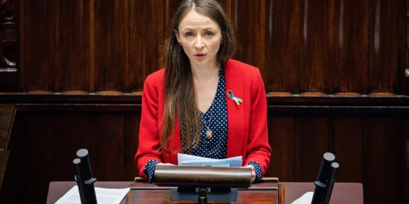 Kobieta w czerwonym żakiecie przemawia z mównicy w sejmie, trzyma kartki, a na klapie ma niebiesko-żółtą wstążkę.