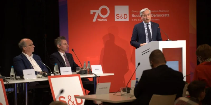 Mężczyzna w garniturze przemawia z mównicy podczas panelu konferencyjnego, na tle czerwonego banera z logo S&D i napisem „70 Years”, przy stole siedzą inni paneliści.