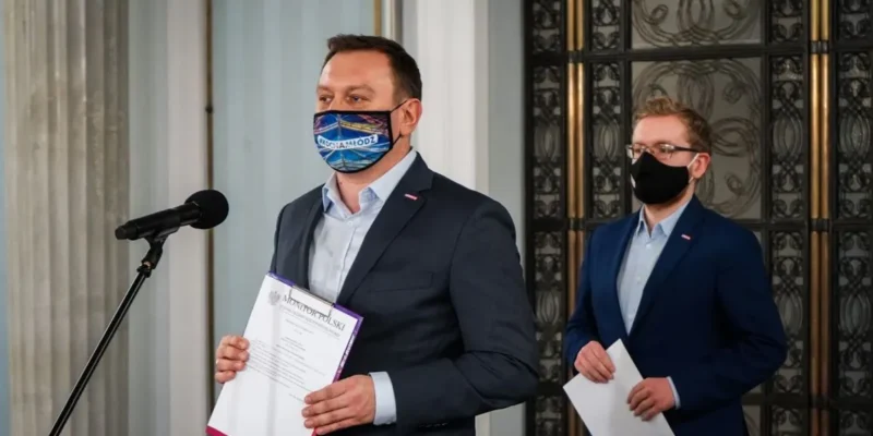 Dwóch mężczyzn w garniturach i maseczkach podczas formalnego wydarzenia; jeden przemawia do mikrofonu i trzyma dokument, drugi stoi z kartką w dłoni, a za nimi widoczne ozdobne drzwi.