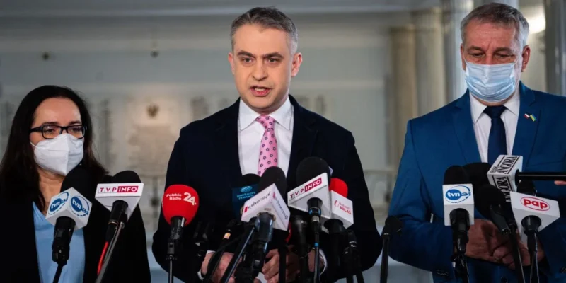 Na zdjęciu trzej mówiący razem politycy podczas konferencji prasowej, otoczeni mikrofonami z różnych mediów.