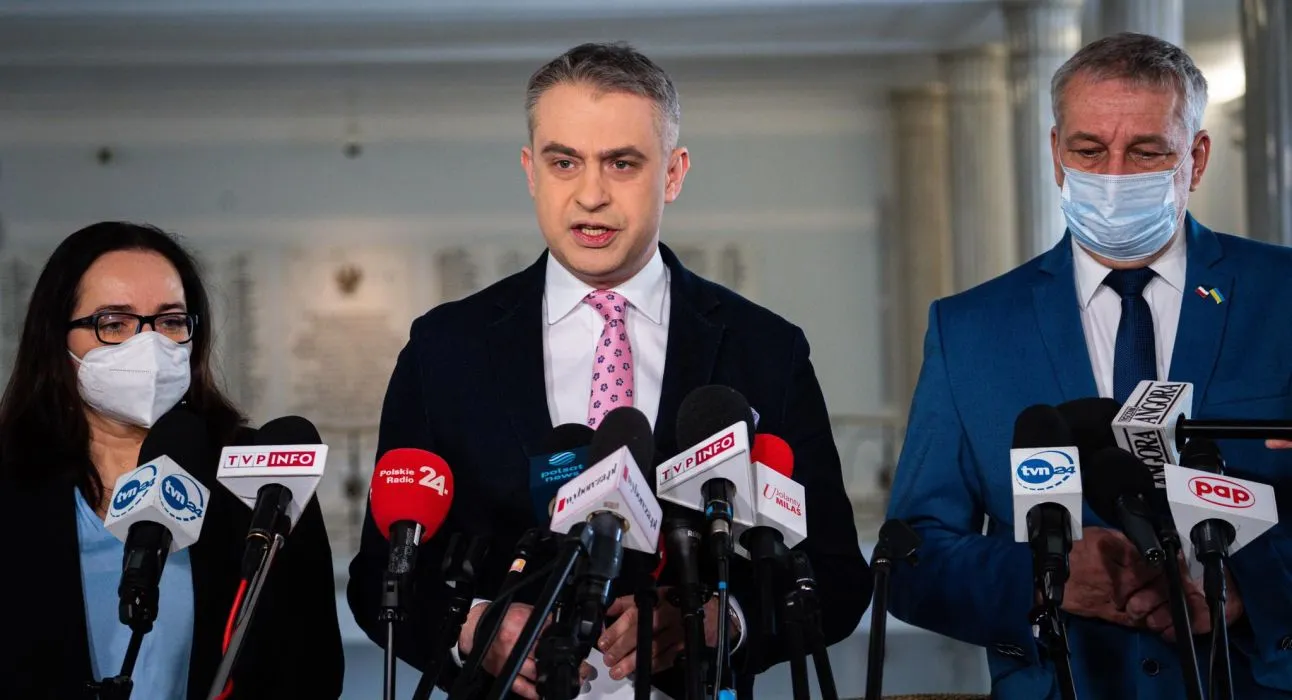 Na zdjęciu trzej mówiący razem politycy podczas konferencji prasowej, otoczeni mikrofonami z różnych mediów.