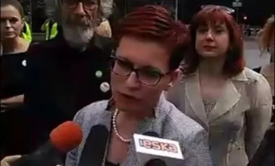 Kobieta w okularach udziela wywiadu przed mikrofonami, a za nią stoją inne osoby podczas wydarzenia Komitet Ratujmy Kobiety (SLD).