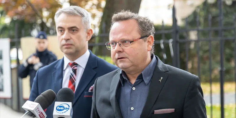 Dwóch mężczyzn w garniturach udziela konferencji prasowej na zewnątrz, stojąc przed mikrofonami i ogrodzeniem w tle.