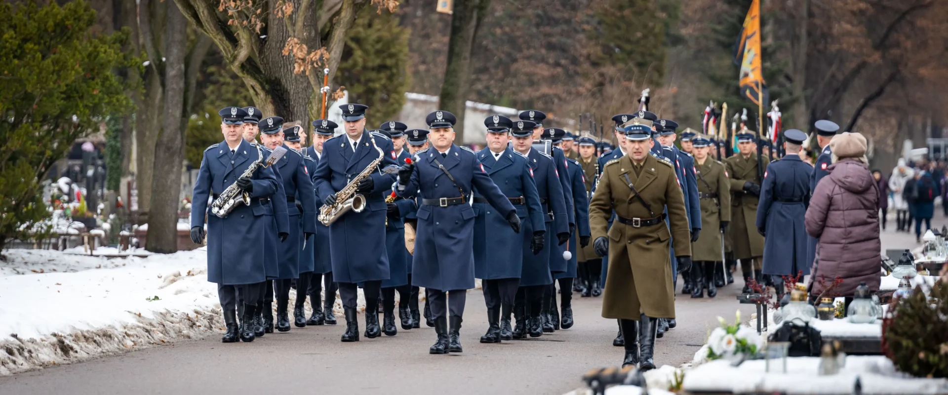 Na zdjęciu widać mundurowych mężczyzn i kobiet w czasie ceremonii, maszerujących cmentarną alejką w zimowej scenerii.