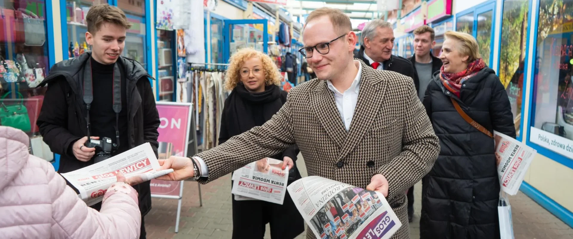 Grupa ludzi na targowisku rozdaje gazety, a stojący na pierwszym planie mężczyzna w kraciastym płaszczu wręcza jedną z nich przechodniom.