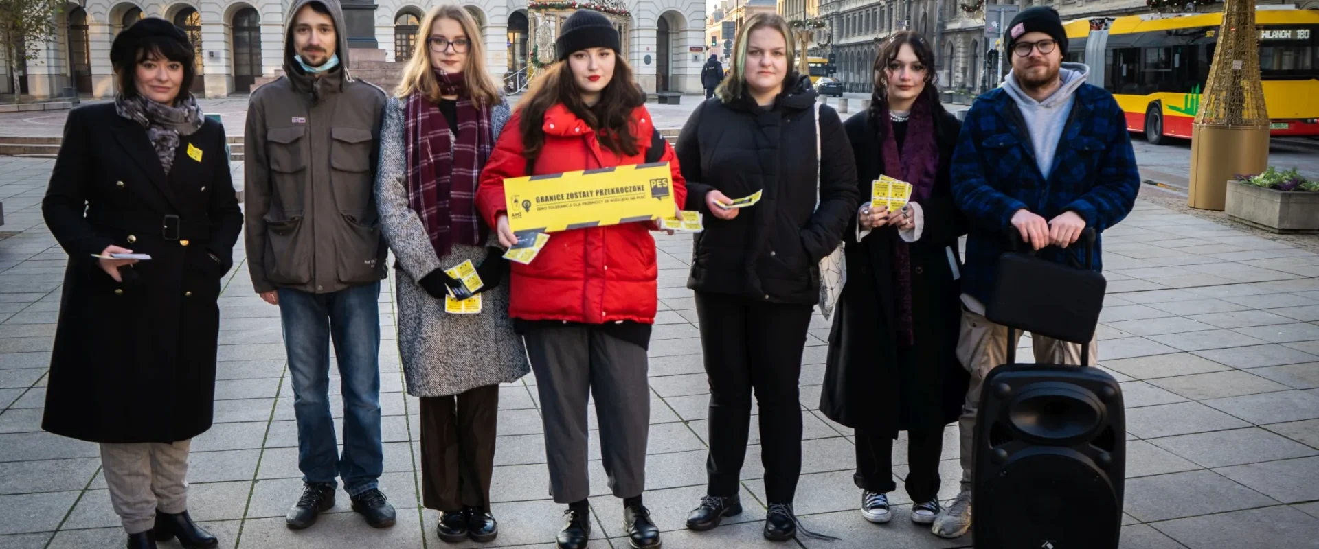 Grupa osób stoi na placu, trzymając transparenty i ulotki, w tle widoczne budynki i pojazdy komunikacji miejskiej.