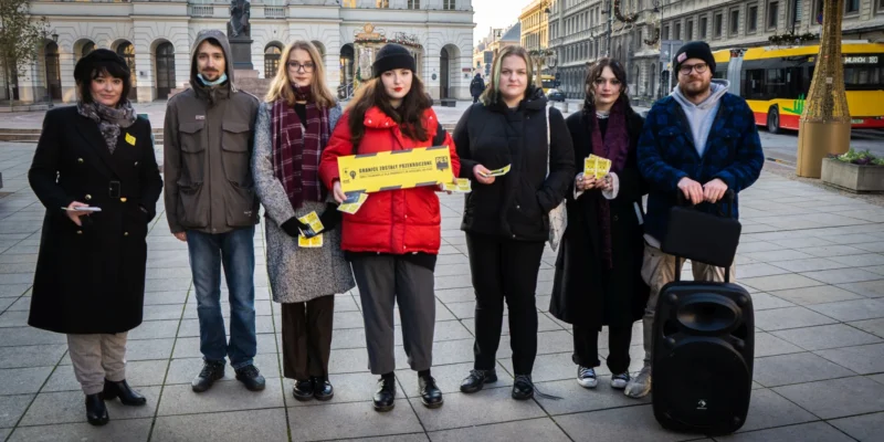 Grupa osób stoi na placu, trzymając transparenty i ulotki, w tle widoczne budynki i pojazdy komunikacji miejskiej.