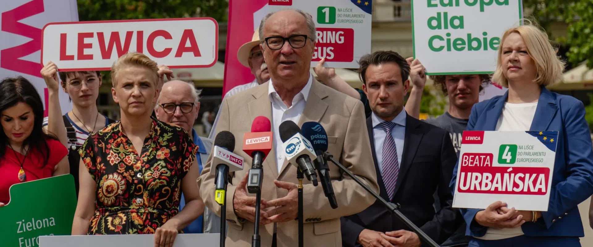 Polityk Lewicy przemawia na konferencji prasowej, otoczony zwolennikami i plakatami Lewicy promującymi wzmocnienie UE i bezpieczeństwa Polski.