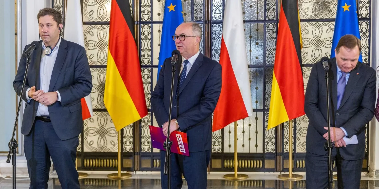 Trzech mężczyzn, w tym jeden trzymający dokumenty, stoi przy mikrofonach w otoczeniu flag niemieckich, polskich i unijnych.