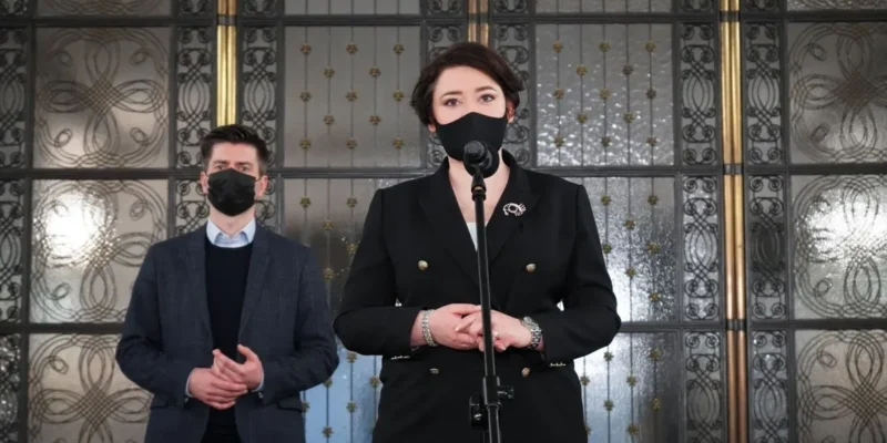Kobieta w czarnym garniturze stoi przy mikrofonie w maseczce, obok niej mężczyzna w marynarce także w maseczce, na tle ozdobnych metalowych drzwi.