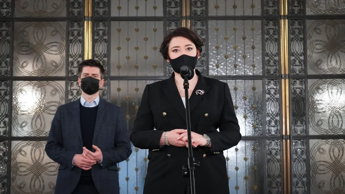 Kobieta w czarnym garniturze stoi przy mikrofonie w maseczce, obok niej mężczyzna w marynarce także w maseczce, na tle ozdobnych metalowych drzwi.