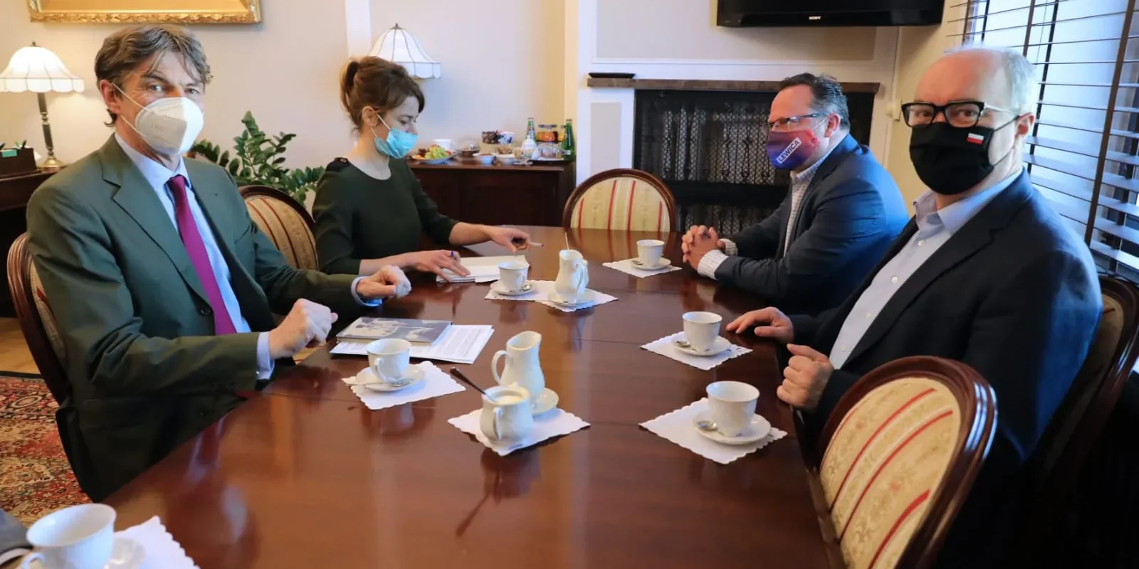 Czterech polityków w maseczkach siedzi przy prostokątnym stole w Sejmie podczas spotkania z przedstawicielem Niemiec; na stole są filiżanki z kawą, a w tle widać obraz i kominek.