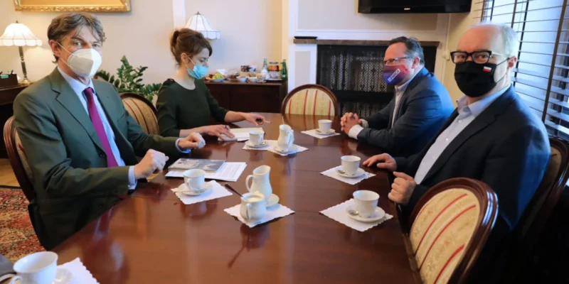 Czterech polityków w maseczkach siedzi przy prostokątnym stole w Sejmie podczas spotkania z przedstawicielem Niemiec; na stole są filiżanki z kawą, a w tle widać obraz i kominek.