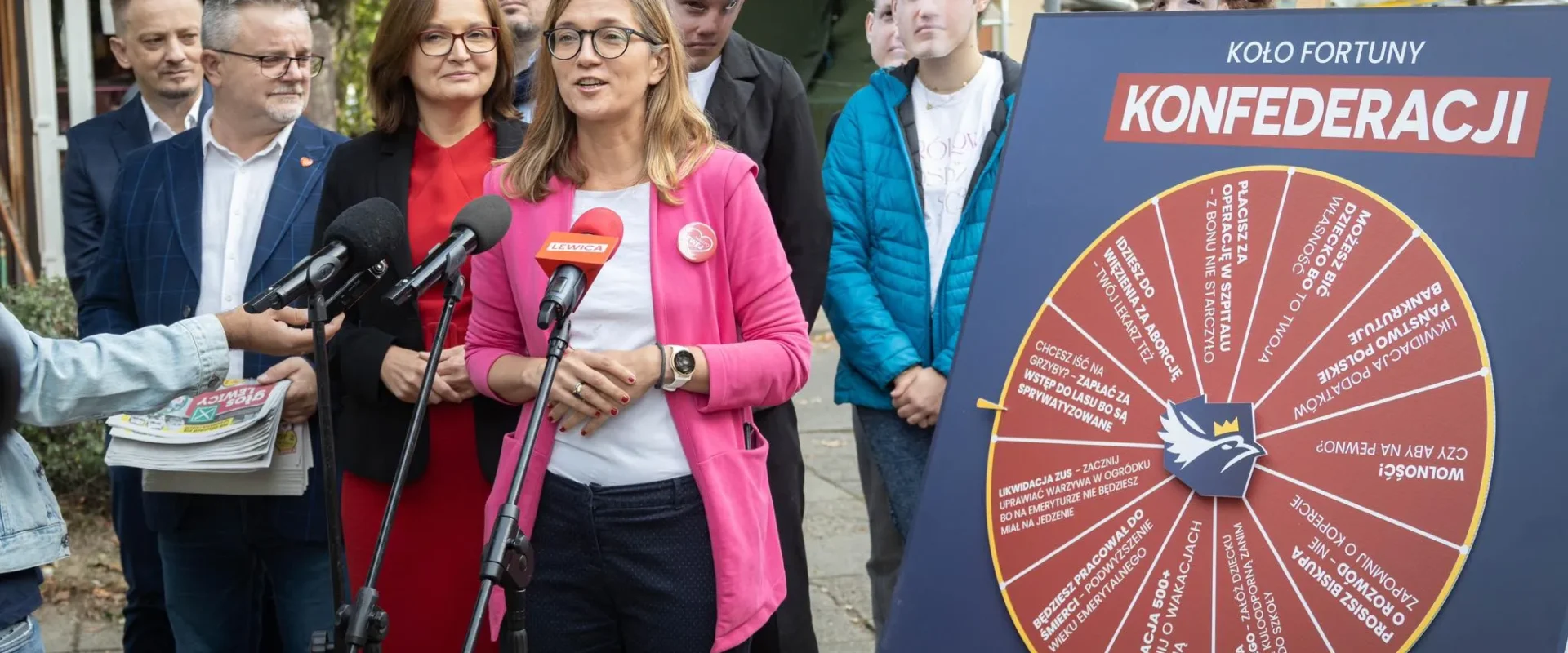 Grupa osób na konferencji prasowej stoi obok dużego koła fortuny z napisem „KOŁO FORTUNY KONFEDERACJI” ustawionego na stojaku.