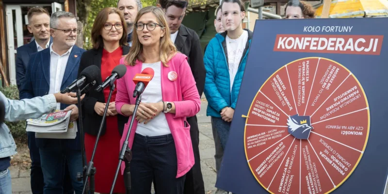 Grupa osób na konferencji prasowej stoi obok dużego koła fortuny z napisem „KOŁO FORTUNY KONFEDERACJI” ustawionego na stojaku.