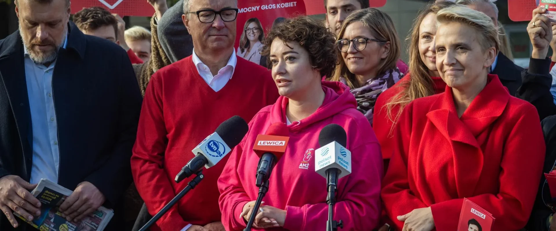 Grupa polityków Lewicy podczas konferencji prasowej, z kobietą w różowej bluzie mówiącą do mikrofonów, otoczona plakatami i innymi członkami ugrupowania.