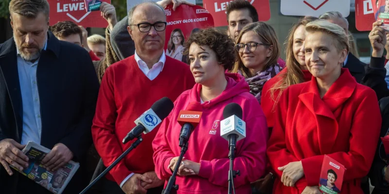 Grupa polityków Lewicy podczas konferencji prasowej, z kobietą w różowej bluzie mówiącą do mikrofonów, otoczona plakatami i innymi członkami ugrupowania.