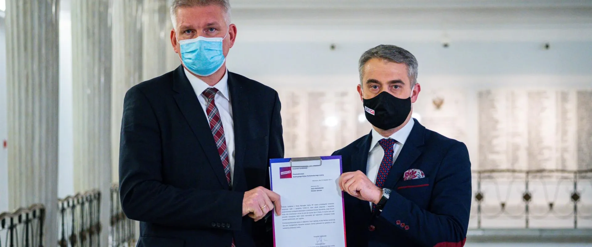 Dwóch mężczyzn w maseczkach, stojących w jasnym wnętrzu, trzymających dokument w rękach.