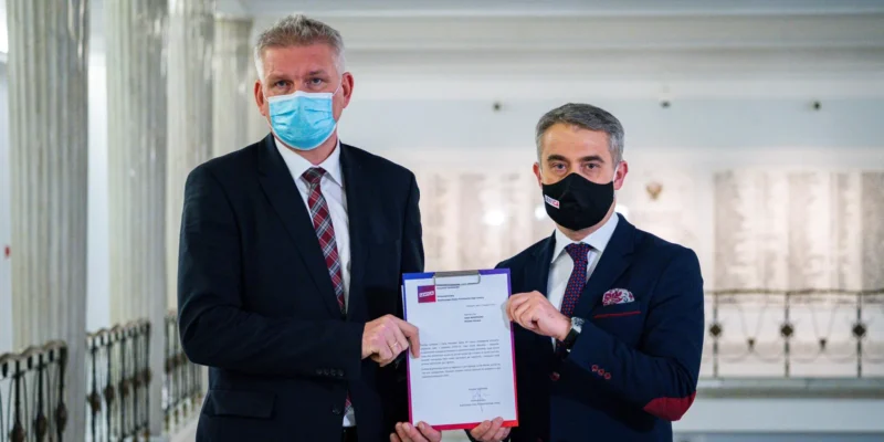 Dwóch mężczyzn w maseczkach, stojących w jasnym wnętrzu, trzymających dokument w rękach.