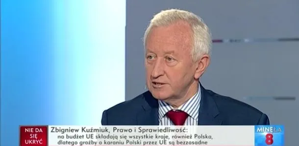 Starszy mężczyzna w garniturze i czerwonym krawacie mówi w studiu telewizyjnym, z paskiem informacyjnym widocznym na dole ekranu.