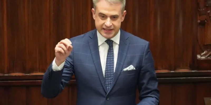 Mężczyzna w granatowym garniturze stoi przy mównicy w sali plenarnej z drewnianymi panelami, gestyklując dłonią.