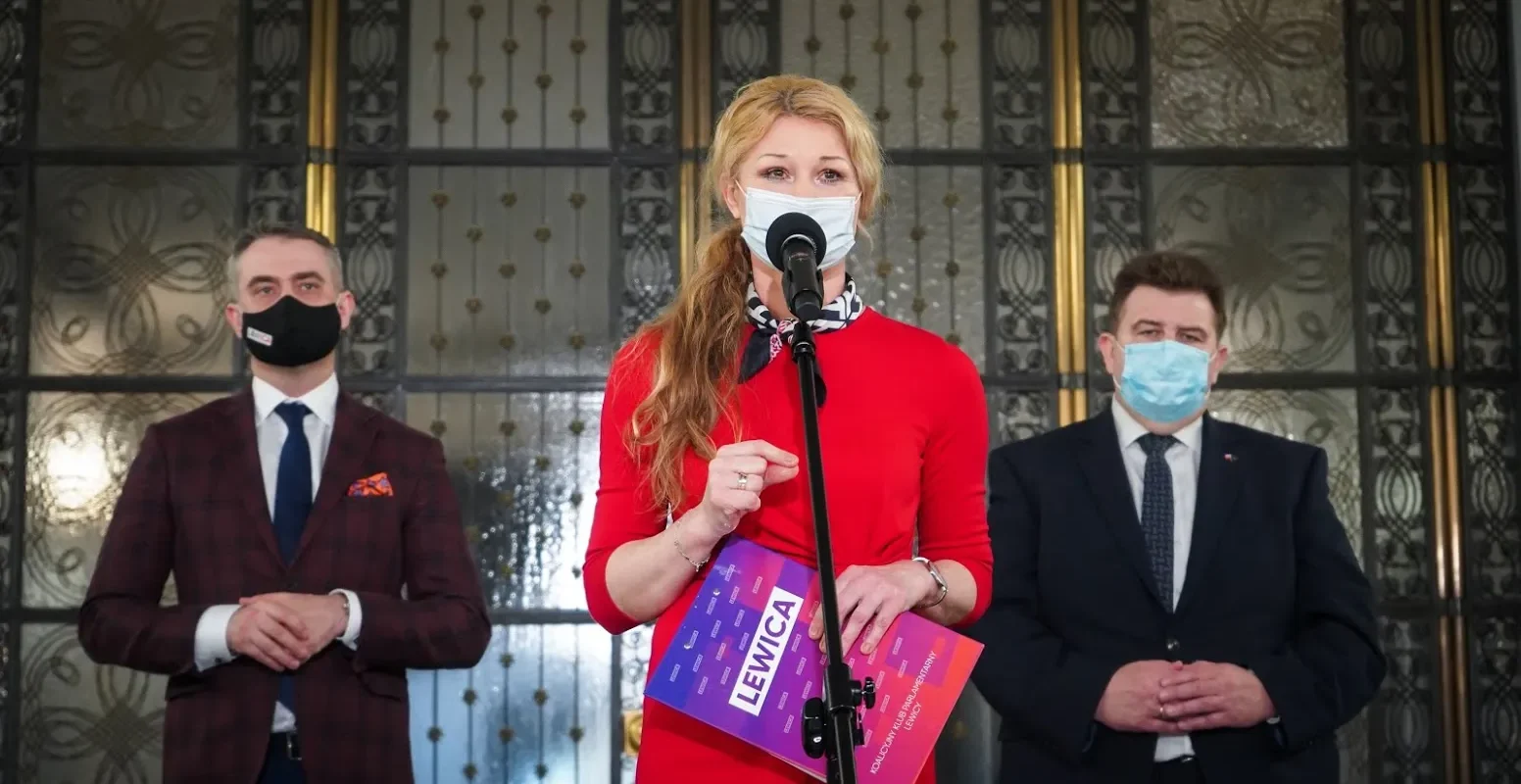 Polityczka w czerwonej sukience przemawia do mikrofonu podczas konferencji prasowej, stojąca między dwoma mężczyznami w garniturach i maseczkach, na tle ozdobnej przeszklonej fasady.