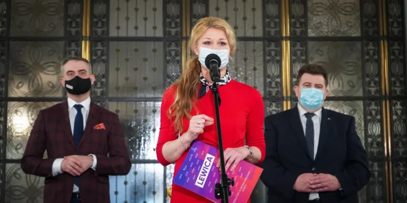 Polityczka w czerwonej sukience przemawia do mikrofonu podczas konferencji prasowej, stojąca między dwoma mężczyznami w garniturach i maseczkach, na tle ozdobnej przeszklonej fasady.