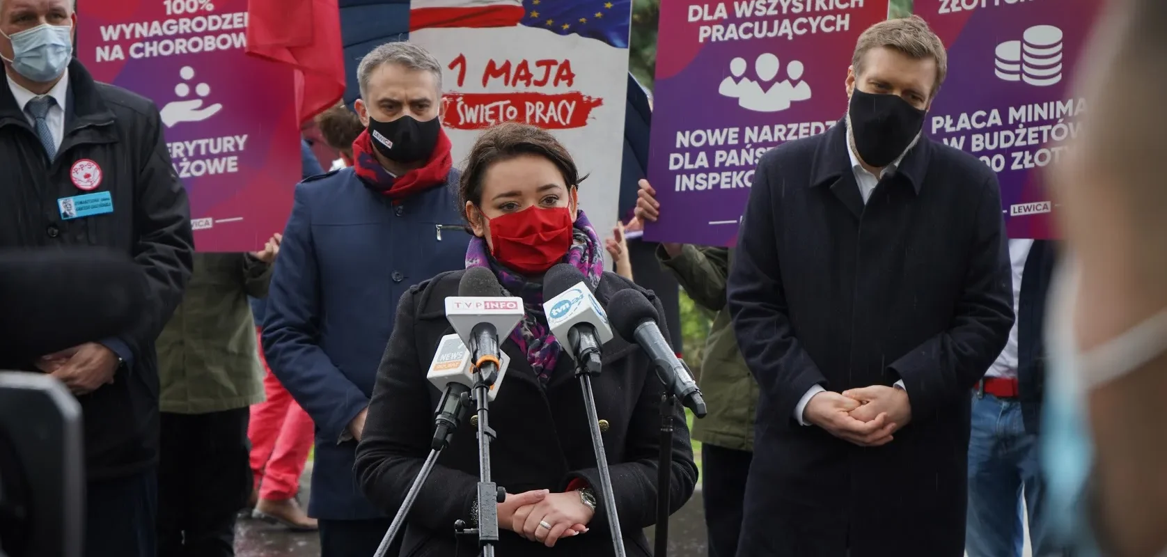 Kobieta w czerwonej masce i ciemnym płaszu przemawia do mikrofonów podczas konferencji prasowej Lewicy, stojąc na tle plakatów i zgromadzonych ludzi.