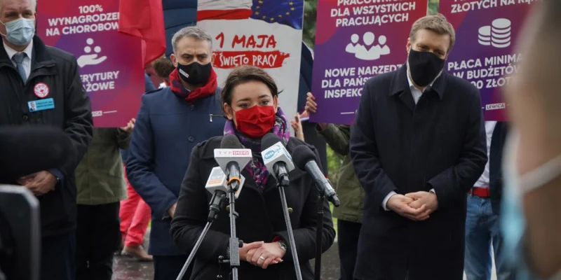 Kobieta w czerwonej masce i ciemnym płaszu przemawia do mikrofonów podczas konferencji prasowej Lewicy, stojąc na tle plakatów i zgromadzonych ludzi.