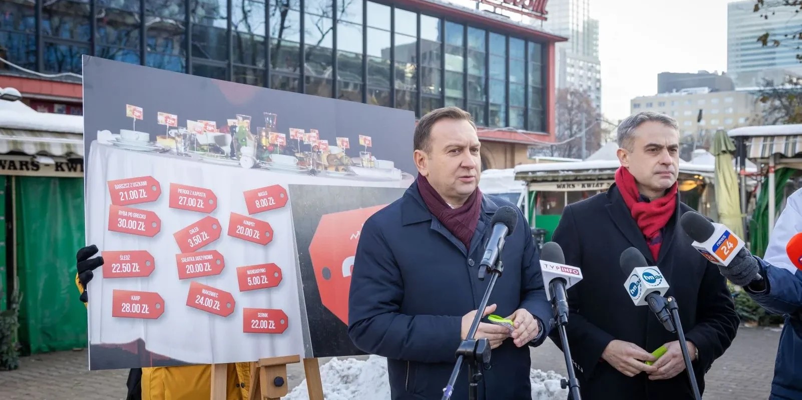 Na zdjęciu dwaj mężczyźni przemawiają przy mikrofonach, obok nich stoi plakat z cenami potraw, a w tle widać budynki i zimową scenerię.