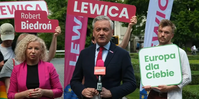 Osoby na konferencji prasowej trzymają banery z napisami Lewica i Europa dla Ciebie, podczas gdy mężczyzna w garniturze przemawia do mikrofonu.