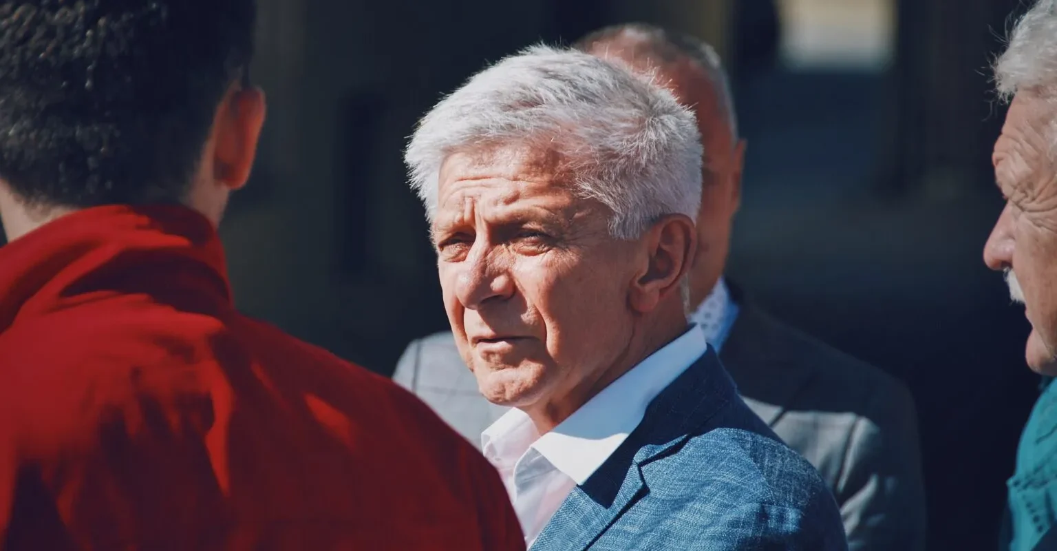 Starszy mężczyzna o siwych włosach w jasnoniebieskim garniturze i białej koszuli stoi na zewnątrz wśród innych osób, patrząc na bok.