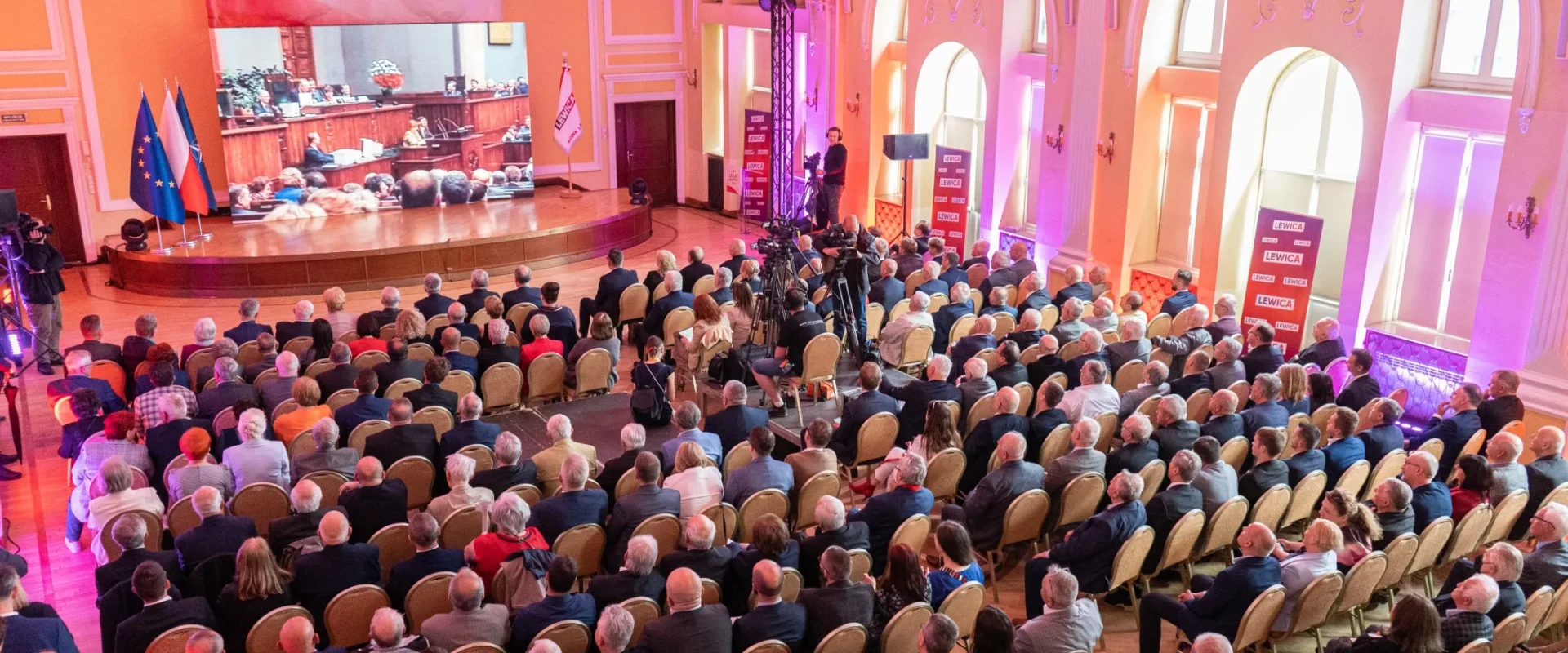 Duża sala konferencyjna wypełniona tłumem uczestników, na scenie prezentacja z flagami UE i Polski i ukazującą 25. rocznicę Konstytucji RP oraz uchwałę Rady Krajowej Nowej Lewicy.