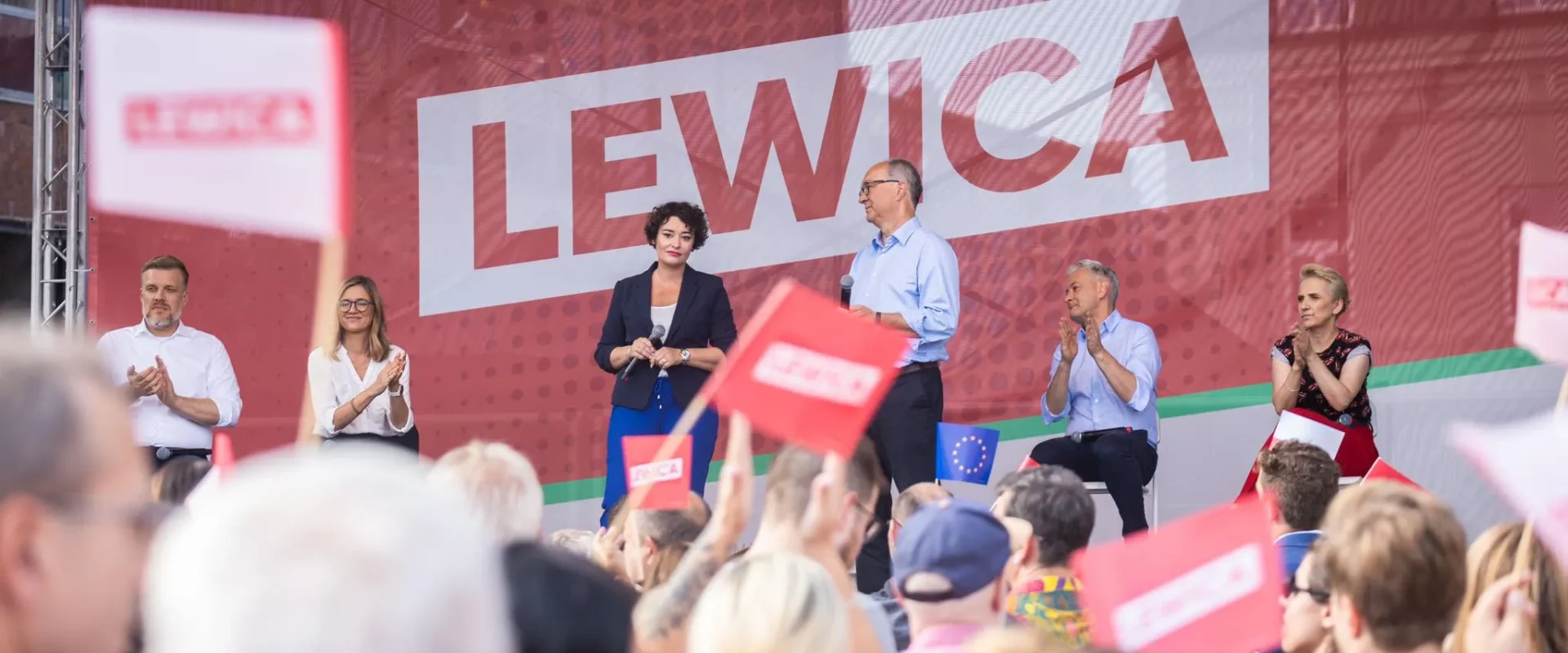 Grupa polityków Lewicy na scenie wiecu w Częstochowie, przed dużą grafiką LEWICA w tle, a tłum widzów trzyma flagi i oklaskuje.