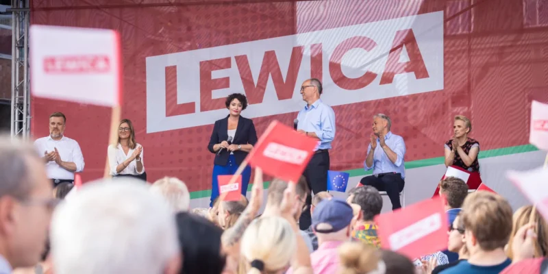 Grupa polityków Lewicy na scenie wiecu w Częstochowie, przed dużą grafiką LEWICA w tle, a tłum widzów trzyma flagi i oklaskuje.